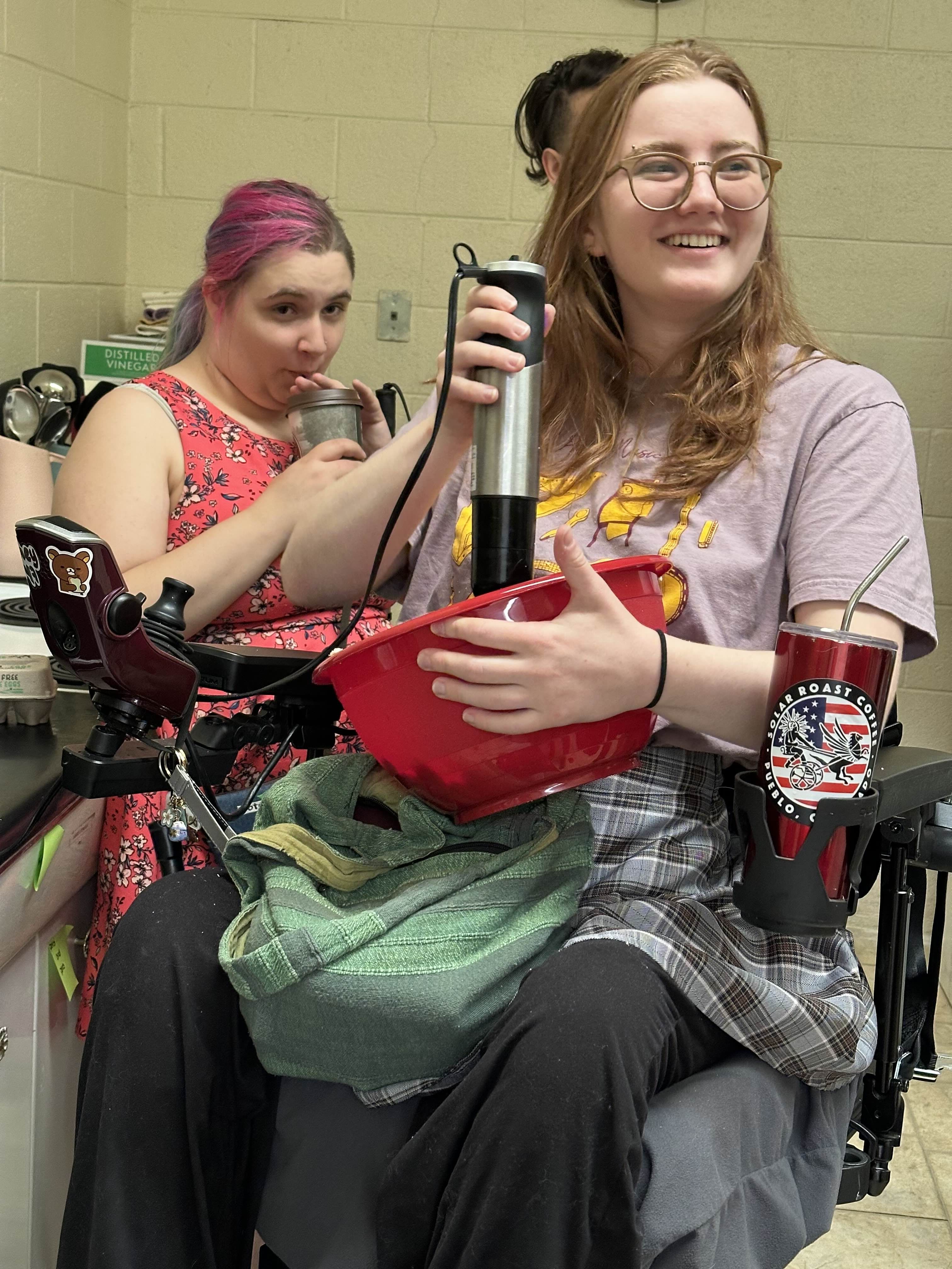 2 people in a kitchen. One is drinking from a straw the other is smiling and mixing something in a bowl